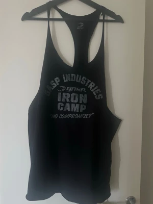 Svart linnetopp från GASP Industries - Säljer en svart linnetopp från GASP Industries med trycket 'IRON CAMP' och 'NO COMPROMISES' framtill. Modellen har djupa ärmhål och brottarrygg, perfekt för gymmet eller träning. Materialet är mjukt och lätt, vilket ger en skön känsla under träningspasset.