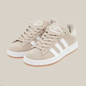 Beige Adidas sneakers med vita ränder - Snygga beige sneakers från Adidas med klassiska vita ränder på sidorna och Adidas-logga på plösen. Skorna har en låg silhuett, snörning och en vit sula med gummidetalj. Ovandelen är i mockaliknande material för en clean och stilren look.
