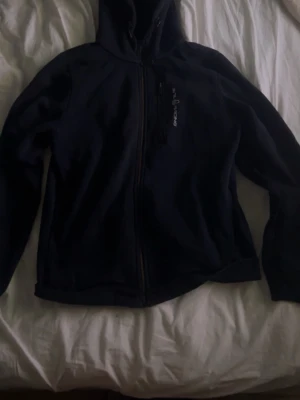 Mörkblå hoodie från Sail Racing - Mörk, stilren hoodie från Sail Racing med dragkedja framtill och en bröstficka med dragkedja. Loggan är tryckt på bröstet. Hoodien har huva och är tillverkad i ett mjukt material som känns skönt mot huden. Perfekt för en avslappnad look.