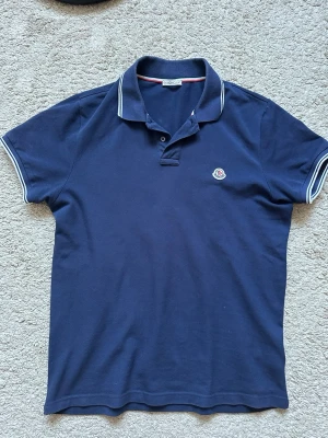 Marinblå pikétröja från Moncler - Snygg marinblå pikétröja från Moncler med vitrandiga detaljer på krage och ärmslut. Klassisk passform och liten Moncler-logga på bröstet. Perfekt till jeans eller shorts för en clean och stilren look.