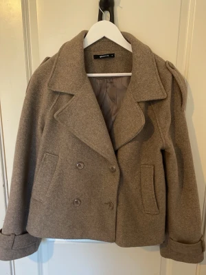 Jacka Gina tricot  - Kort beige rock från Gina Tricot med dubbelknäppning och bred krage. Rocken har två framfickor och uppvikta ärmslut. Tillverkad i ett ull-liknande material med slät insida. Perfekt för kyliga dagar och ger en stilren look. I storlek M 