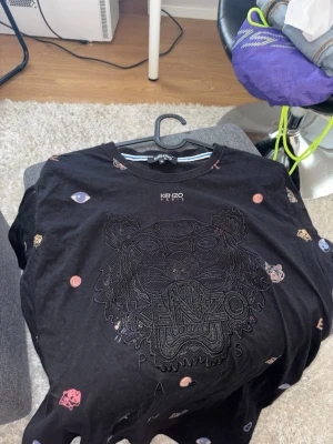 Svart Kenzo t-shirt med tigerbrodyr - Kenzo t-shirt i svart bomull med ikonisk tigerbrodyr framtill och färgglada små broderade symboler över hela plagget. Klassisk passform och rund hals. Perfekt för dig som vill ha en unik och trendig look.