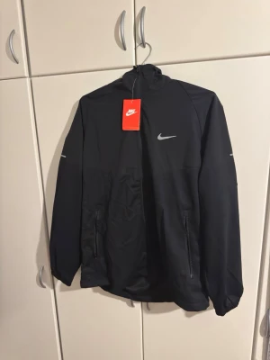 Svart Nike vindjacka med huva - Snygg svart vindjacka från Nike med reflekterande logga på bröstet och ryggen. Jackan har dragkedjefickor, hög krage och huva. Tillverkad i lätt och tåligt material, perfekt för blåsiga dagar. Sportig design med elastiska muddar och diskreta detaljer.OBS STORLEK PÅ TAGG SÄGER XXL MEN PASSAR EGENTLIGEN M-L