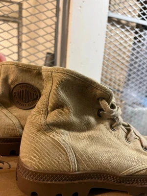 Beige canvas kängor från Palladium - Säljer ett par beige kängor från Palladium i slitstark canvas med bruna gummisulor och tåhätta. Klassisk hög modell med snörning och Palladium-logga på sidan och plösen. Perfekta för dig som gillar streetwear och vill ha något bekvämt och stilrent.