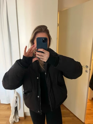 Svart pufferjacka med dragkedja - Säljer en svart pufferjacka med dragkedja och tryckknappar. Jackan har två stora fickor framtill och en hög krage. Lite oversized passform men lite kortare modell. Använd men i mycker bra skick! 