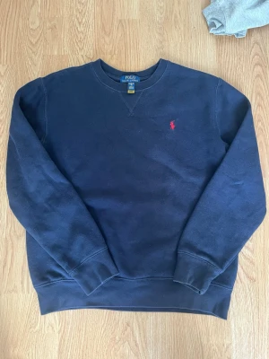 Mörkblå sweatshirt från Polo Ralph Lauren - Klassisk mörkblå sweatshirt från Polo Ralph Lauren med rund halsringning och röd broderad logga på bröstet. Tröjan har ribbade muddar vid ärmslut och nederkant. Perfekt för en avslappnad och stilren look. Storlek XS! Skriv till mig vid intresse eller frågor! MVH 🤗