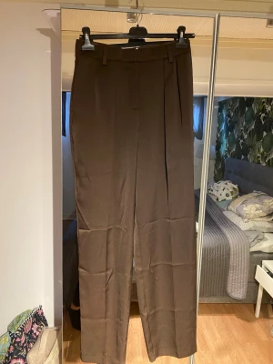 Mörkbruna nya kostymbyxor från Primark stl 38 - Snygga bruna något glansiga kostymbyxor från Primark med raka ben och normalhög midja. Byxorna har bälteshällor och pressveck framtill för en clean look. Perfekta att styla med en skjorta eller crop top för en trendig vibe.