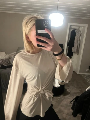 Beige topp med knyt i midjan - Trendig beige topp med långa ärmar och snygg knytning i midjan. Toppen har en rund halsringning och är tillverkad i ett mjukt material som ger en relaxed men ändå stilren look. Perfekt att matcha med jeans eller kjol för en cool outfit.