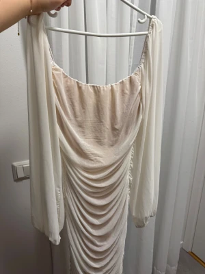 Vit långklänning med rynkade detaljer - Elegant vit långklänning med rynkade detaljer framtill och baktill. Klänningen har långa, lösa ärmar och en bred, rundad urringning. Tillverkad i ett lätt, flytande material som ger en romantisk känsla. Perfekt för speciella tillfällen.