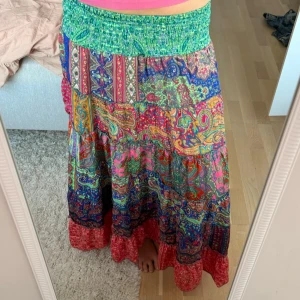 Färgglad långkjol med paisleymönster - Säljer en bohemisk långkjol med elastisk grön midja och färgstarkt paisleymönster i rosa, blått, grönt och rött. Kjolen har volangdetaljer och är perfekt för dig som gillar statement-plagg och sköna material.