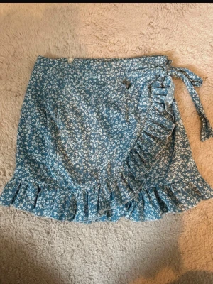 Blommig blå omlottkjol med volang - Supersöt blå kort omlottkjol med vitt blommönster och volangkant. Kjolen har knytband i midjan och är perfekt för varma dagar. Lätt och luftig känsla, passar till både sneakers och sandaler.