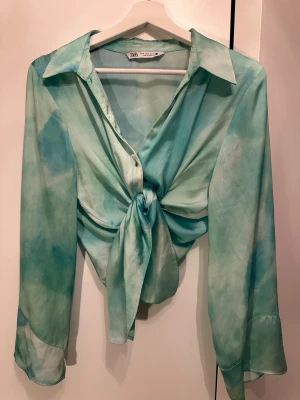 Grönblå knytblus från Zara - Säljer en grönblå skjorta från Zara i ett tunt, glansigt material. Skjortan har långa ärmar, klassisk krage och kan knytas framtill för en trendig look. Perfekt för dig som gillar färg och vill sticka ut lite extra.