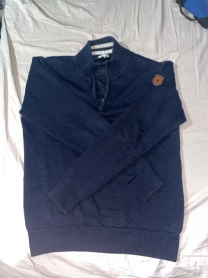 Mörkblå stickad tröja med halvzip - Mörkblå stickad tröja från Marine storlek M med halvzip och ståkrage. Tröjan har ribbade muddar vid ärmslut och nederkant samt en snygg läderpatch med logga på axeln. Perfekt för lager-på-lager och chill stil.