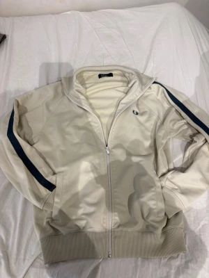Beige track jacket från Fred Perry - Snygg beige track jacket från Fred Perry med marinblå ränder längs ärmarna och dragkedja framtill. Jackan har ribbade muddar, ståkrage och två fickor. Perfekt för en sportig och avslappnad stil.