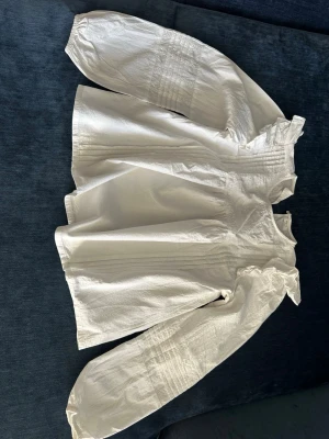 Vit blus med volangdetaljer - Söt vit blus med volangdetaljer vid axlarna och rynkade, vida långärmar. Blusen har en lös passform och fina sömdetaljer framtill och på ärmarna. Tillverkad i ett lätt, vävt material som ger en romantisk och trendig look.