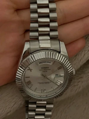 Silverfärgad klocka från NIBOSI - Stilren klocka i rostfritt stål från NIBOSI med silverfärgad länkarmband och urtavla. Klockan har dag- och datumvisning, romerska siffror och räfflad bezel. Perfekt accessoar för dig som gillar klassisk och tidlös design.