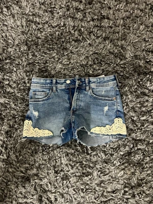 Blå jeansshorts med spetsdetaljer - Säljer ett par blå jeansshorts med rå kant och slitningar. Shortsen har vita spetsdetaljer på båda benen och klassisk femficksdesign. Perfekta för sommaren och ger en avslappnad vibe.