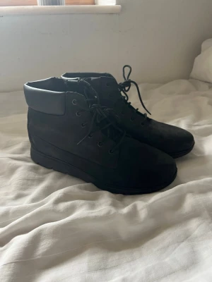 Svarta kängor med snörning - Säljer ett par svarta kängor från Timberland med snörning och vadderad kant runt ankeln. De har en stilren design och är tillverkade i mockaliknande material. 