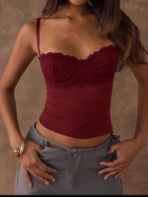 Vinröd spetstopp med tunna axelband - Snygg vinröd topp med tunna justerbara axelband och vacker spetsdetalj över bysten. Toppen har en figurnära passform, lätt transparent tyg och öppen rygg med spänne. Perfekt för att skapa en trendig och feminin look. Storlek S!