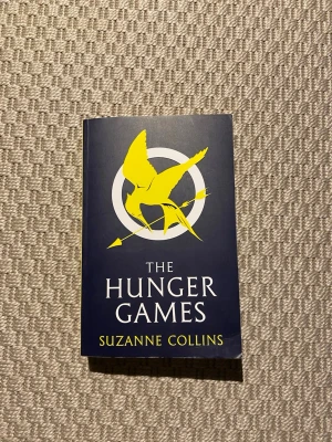 The Hunger Games pocketbok - Säljer boken The Hunger Games av Suzanne Collins. Omslaget är mörkblått med en stor gul fågel och vit text. Baksidan är gul med svart text. Perfekt för dig som gillar spänning och dystopiska berättelser.