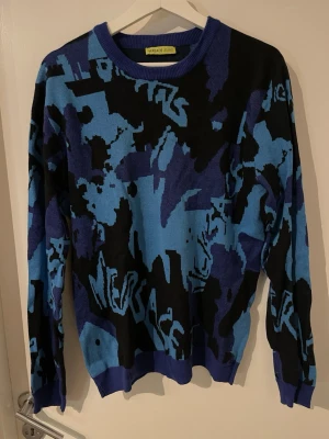 Blå och svart stickad tröja Versace Jeans - Cool stickad tröja från Versace Jeans i blått och svart med grafiskt mönster och textdetaljer. Tröjan har rund halsringning, långa ärmar och ribbade muddar. OBS strlk officiellt XL men sitter som M/L. 