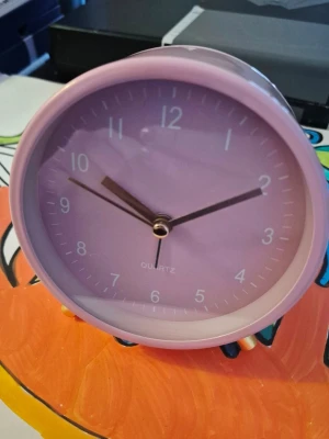 Rund rosa väckarklocka med bakgrundsbelysning.  - Säljer en stilren rund växkarklocka i ljusrosa med matchande urtavla och vita siffror. Klockan har tre visare i metall och står på små guldfärgade ben. Perfekt som dekoration eller för att hålla koll på tiden i rummet. Klockan är cirka 11 cm i diameter plus fötter