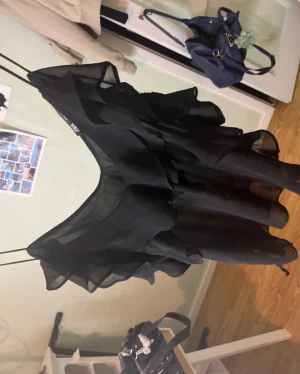 Svart volangklänning från Zara - Supersnygg svart kortklänning från Zara med volangdetaljer och tunn, luftig känsla. Klänningen är ärmlös och har en feminin siluett som ger en chic vibe. Perfekt för dig som vill sticka ut med en trendig och elegant look.