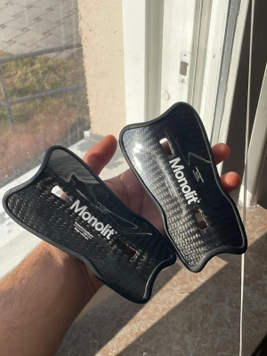 Monolit benskydd  - Säljer ett par svarta benskydd från Monolit, tillverkade i kolfiber med snygg glansig finish och ergonomisk form. Perfekta för dig som vill ha lätta och slitstarka benskydd till fotbollsträningen eller matchen. Dem små!