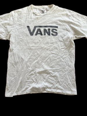 Vit Vans t-shirt med logga - Klassisk vit t-shirt från Vans med stor svart logga tryckt på bröstet. T-shirten har rund halsringning och korta ärmar. Tillverkad i mjuk bomull som känns skön mot huden. Perfekt för en avslappnad streetwear-look.