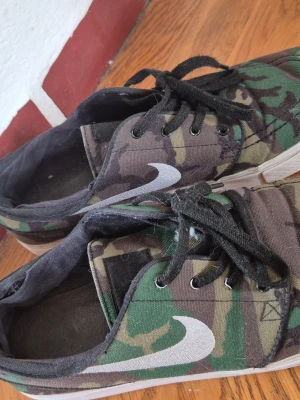 Nike camo militärmönster sneakers med svart snörning - Nike sneakers med camouflagemönster / militärmönster i grönt, brunt och svart. Skorna har svart snörning, vit sula och den klassiska vita Nike-loggan på sidan. Ovandelen är i textil och insidan är svart. Är i väldigt bra skick på utsidan men lite slitna i. Använts få gånger ute men en del inne då de är lite små och därför har min kille försökt töja ut dem. Se bild för hål på insidan häl, häl i båda skorna är mest slitna.