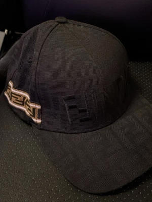 Svart keps från Fendi med logga - Snygg svart keps från Fendi med ton-i-ton Fendi-mönster över hela kepsen och en broderad logga framtill. På sidan finns ett band med Fendi-loggan i beige och svart. Tillverkad i Italien och har en klassisk böjd skärm. Perfekt för dig som gillar exklusiva accessoarer.