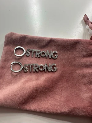 IOAKU örhängen strong - Säljer ett par statement-örhängen från IOAKU i silverfärg med ordet STRONG i stora bokstäver täckta av gnistrande strass. ny pris 599kr