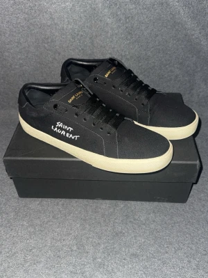 Saint Laurent svarta låga sneakers - Svarta sneakers från Saint Laurent med vit sula och handskriven logga på sidan. Skorna har klassisk låg profil, svarta skosnören och ovandel i canvas. Guldtext på plösen ger en lyxig touch. Perfekta för dig som gillar stilrena och exklusiva sneakers.