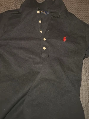Svart pikétröja från Ralph Lauren - Klassisk svart pikétröja från Ralph Lauren med vit knappslå och röd broderad logga på bröstet. Tröjan har krage och korta ärmar, perfekt för en stilren look. Tillverkad i bomull för skön komfort.