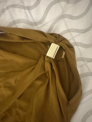 Brun oneshoulder topp med metallspänne - Snygg brun oneshoulder topp med ett coolt metallspänne på axeln. Toppen har en asymmetrisk design och är gjord i ett mjukt, stretchigt material som sitter skönt på kroppen. Perfekt för dig som vill sticka ut lite extra.