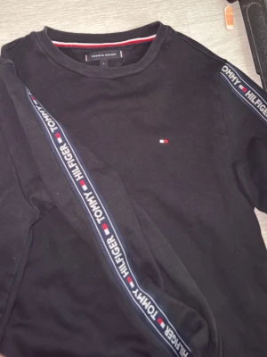Svart sweatshirt Tommy Hilfiger - Svart sweatshirt från Tommy Hilfiger med rund halsringning och ribbade muddar. Tröjan har logoband längs ärmarna och det klassiska lilla Hilfiger-flaggan broderat på bröstet. Perfekt för en avslappnad och stilren look.