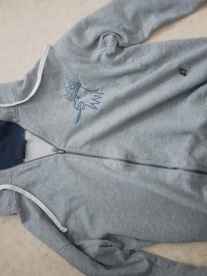 Ljusgrå hoodie med dragkedja - Ljusgrå hoodie från Koppartrans med dragkedja framtill och vit kant längs huvan. Broderad Koppartrand-logga med krona på bröstet. Tillverkad i mjuk bomullsblandning och har långa ärmar samt ribbade muddar. Perfekt för en avslappnad och sportig stil.