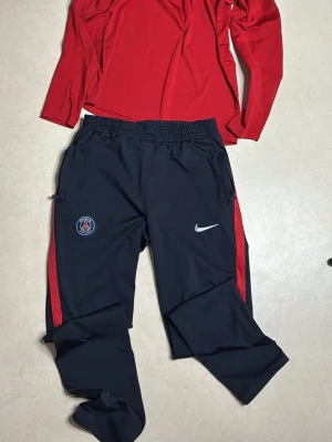 PSG träningsset från Nike röd/svart - Snyggt PSG träningsset från Nike med röd långärmad tröja och svarta byxor med röda detaljer. Tröjan har halv dragkedja och klubbmärke på bröstet. Byxorna har elastisk midja, Nike-logga och PSG-emblem. Perfekt för fotbollsträning eller chill.