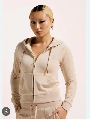 Beige velour hoodie från Juicy Couture - Snygg beige hoodie i mjuk velour från Juicy Couture. Dragkedja framtill, huva med snören och fickor på magen. Ikonisk JC-logga broderad på bröstet. Perfekt för en chill och trendig look.