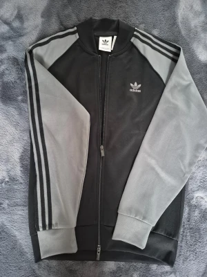 Adidas "Bomber jacka" - Klassisk Adidas track jacket i svart med grå ärmar och svarta ränder längs axlar och armar. Dragkedja framtill, ribbade muddar och Adidas logga på bröstet.  Helt ny,aldrig använd! Nypriset var 800 kr