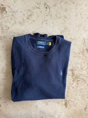 Mörkblå stickad tröja Polo Ralph Lauren - Mörkblå merinoulls tröja från Polo Ralph Lauren med rund halsringning och diskret broderad logga på bröstet. Tröjan har långa ärmar och ribbade muddar vid ärmslut och nederkant. Perfekt för en stilren och klassisk look.