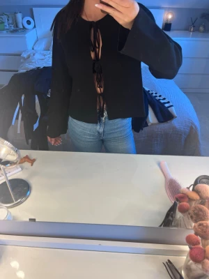 Svart topp med snörning framtill - Trendig svart topp med långa ärmar och snygg snörning framtill som ger en öppen look. Toppen har en loose passform och är perfekt för dig som vill sticka ut. Tillverkad i mjukt material som känns skönt mot huden.