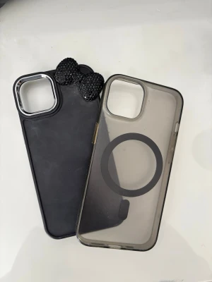 Två mobilskal till iPhone, svart & transparent - Säljer två mobilskal till iPhone 12. Ett svart skal med silverkant runt kameran och dekorerad med svarta glittriga rosetter, samt ett transparent skal med MagSafe-ring. Båda har öppning för kamera och knappar. Perfekt för dig som vill skydda mobilen med stil.