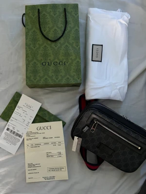 Svart axelväska från Gucci - Säljer en svart axelväska från Gucci med klassiskt GG-mönster och silverfärgad dragkedja. Väskan har en justerbar axelrem i rött och blått samt en praktisk ytterficka med dragkedja. Perfekt för dig som vill ha en stilren och lyxig accessoar.