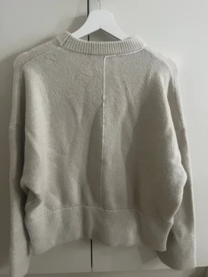 Beige stickad tröja - Säljer en beige  stickad tröja med markerad söm framtill och baktill. Tröjan har bred ribbad mudd vid ärmslut och nederkant samt rund halsringning. Perfekt för lager på lager och en avslappnad look.