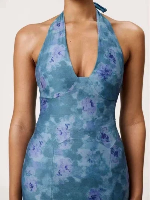 Halterneck klänning Adoore - Säljer en blå maxiklänning med halterneck och djup urringning. Klänningen har ett blommigt mönster i blå och lila toner och är figurnära med lång siluett. Materialet är mjukt och följsamt, perfekt för sommarens alla tillfällen. Den är helt ny, aldrig använd (den passade tyvärr inte). 