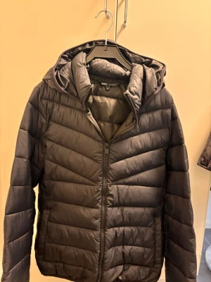 Svart pufferjacka från ONLY med huva - Säljer en svart pufferjacka från ONLY med dragkedja framtill och stor, mysig huva. Jackan har en quiltad design med horisontella sömmar och är perfekt för kalla vinterdagar. Materialet är lätt och ger en sportig look.