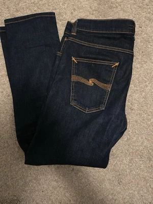 Mörkblå jeans från Nudie Jeans - Lean dean. Säljer ett par mörkblå jeans från Nudie Jeans med klassisk femficksmodell och kontrastsömmar i orange. Jeansen har rak passform och är tillverkade i kraftig denim. Snygg detalj med vågig söm på bakfickan. Perfekta för en clean och stilren look.