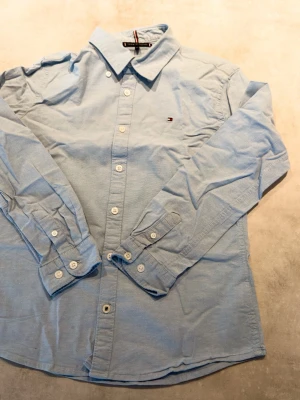 Ljusblå Tommy Hilfiger skjorta - Jag säljer en stilig skjorta från Tommy Hilfiger I barnstorlek M (152), tyvärr har den några riktigt små fläckar (på bilden) men dom uppmärksammas inte riktigt annars är den i riktigt bra skick! Tveka inte att höra av dig, skickas inom en dag!