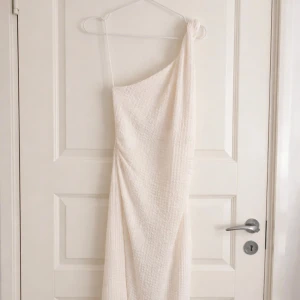 Vit one shoulder klänning - Stilren vit klänning med one shoulder-design och lätt strukturerat tyg från Hm. Klänningen har även en slits vid sidan och etiketten finns kvar då jag aldrig användt den. Dock är klänningen lite genomskinlig. 